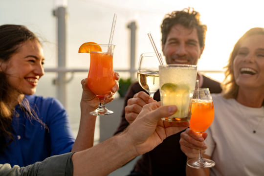 Zwei Frauen, ein Mann stoßen mit Cocktails an auf einem Kreuzfahrtschiff