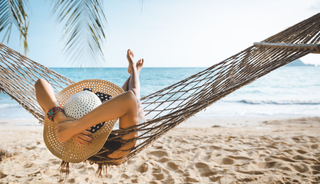 Frau mit Sonnenhut in Hängematte, die Arme hinter dem Kopf verschränkt mit Blick Richtung Meer, unter ihr der feine Sandstrand, oben rechts sind Palemenwedel